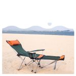 A green and orange camping lounge chair is set up on a sandy beach, facing a calm ocean.