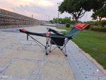 A green and black camping lounge chair with red accents, set on stone paving near grass and trees.