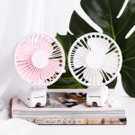 A pink and white rechargeable folding hand-held fan placed on top of a book, against a backdrop of green leaves.