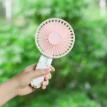 A pink and white rechargeable hand-held fan being held against a green, natural background.