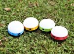 Four portable camping lights in blue, yellow, green, and red, placed on grass. Each light has a circular design.