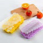 Hexagonal silicone ice cube trays in yellow and purple, with clear lids, on a wooden cutting board alongside fruits.