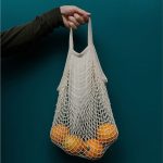 A hand holds a beige cotton mesh bag filled with oranges, set against a dark green background.