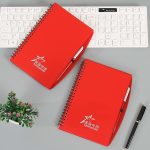 A red spiral notebook with a pen, featuring a logo, placed on a desk with a keyboard and a small plant.