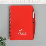 A red spiral notebook with a pen attached, featuring a logo on the front. Ideal for office and business use.