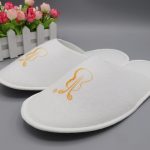 A pair of white disposable hotel slippers with an embroidered design, placed on a grey surface near decorative flowers.