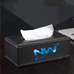 A black leather tissue box with a white tissue emerging from the top. It features a logo on one side.