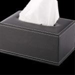 A sleek black leather tissue box with white stitching and a tissue peeking out from the top.