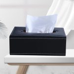 A black leather tissue box with white stitching, featuring a slot for tissues, placed on a light-coloured table.