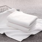 A stack of white bath towels neatly arranged on a soft surface, alongside a wire basket.