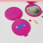 A round, pink folding mirror with a reflective surface and a logo on the outer casing.