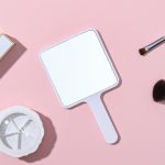 A square handheld mirror with a white handle, placed on a pink background alongside beauty tools.