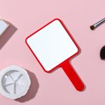 A square handheld mirror with a red frame, placed on a pink surface alongside cosmetic items.