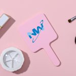 A square handheld mirror in pink with a branded logo, placed on a light pink surface alongside makeup accessories.