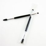 A double-headed eyebrow brush with a black grip, featuring a spoolie on one end and an angled brush on the other, in clear packaging.