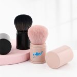 Two retractable makeup brushes in black and pink, featuring soft bristles, displayed on a pastel surface. One has a logo.