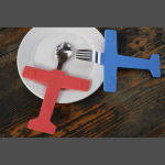A red airplane-shaped spoon and a blue airplane-shaped fork on a white plate against a wooden background.