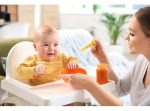 A baby sits in a high chair, smiling at a woman holding a yellow spoon and a jar of orange food.
