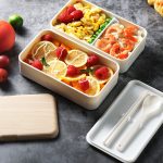 A double-layer lunch box set in white containing fruits, vegetables, scrambled eggs, and seafood, with a wooden lid and utensils.