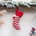 A red and white chevron-patterned Christmas stocking with a quilted top and pom-pom decoration, hanging from a mantel.
