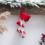 A red and white Christmas stocking decorated with a reindeer pattern and a pom-pom, hanging from a wooden shelf.