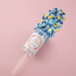A clear container filled with blue and gold confetti, topped with a plastic straw and a label.