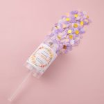 A clear push pop filled with purple, pink, and gold confetti against a pink background, featuring a decorative label and a straw.