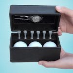 A black golf set box containing three white golf balls, six wooden tees, and a silver divot tool.