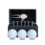 A black box containing three white golf balls, a silver pitchfork tool, and wooden tees. The box has a logo.