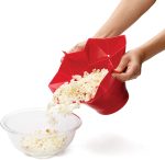A red popcorn bucket spilling popcorn into a glass bowl, featuring a geometric design and held by a hand.