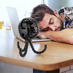 A black stroller fan with a flexible tripod sits on a table beside a person resting their head on a laptop.