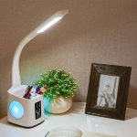 A white LED desk lamp with a flexible neck, USB charging port, digital clock, and a plant beside a photo frame on a desk.