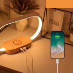 A wooden bedside lamp with a glowing light and a smartphone charging beside it. The lamp has a logo on its base.