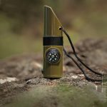 A green camping whistle with a built-in compass, attached to a black lanyard, resting on a rocky surface.
