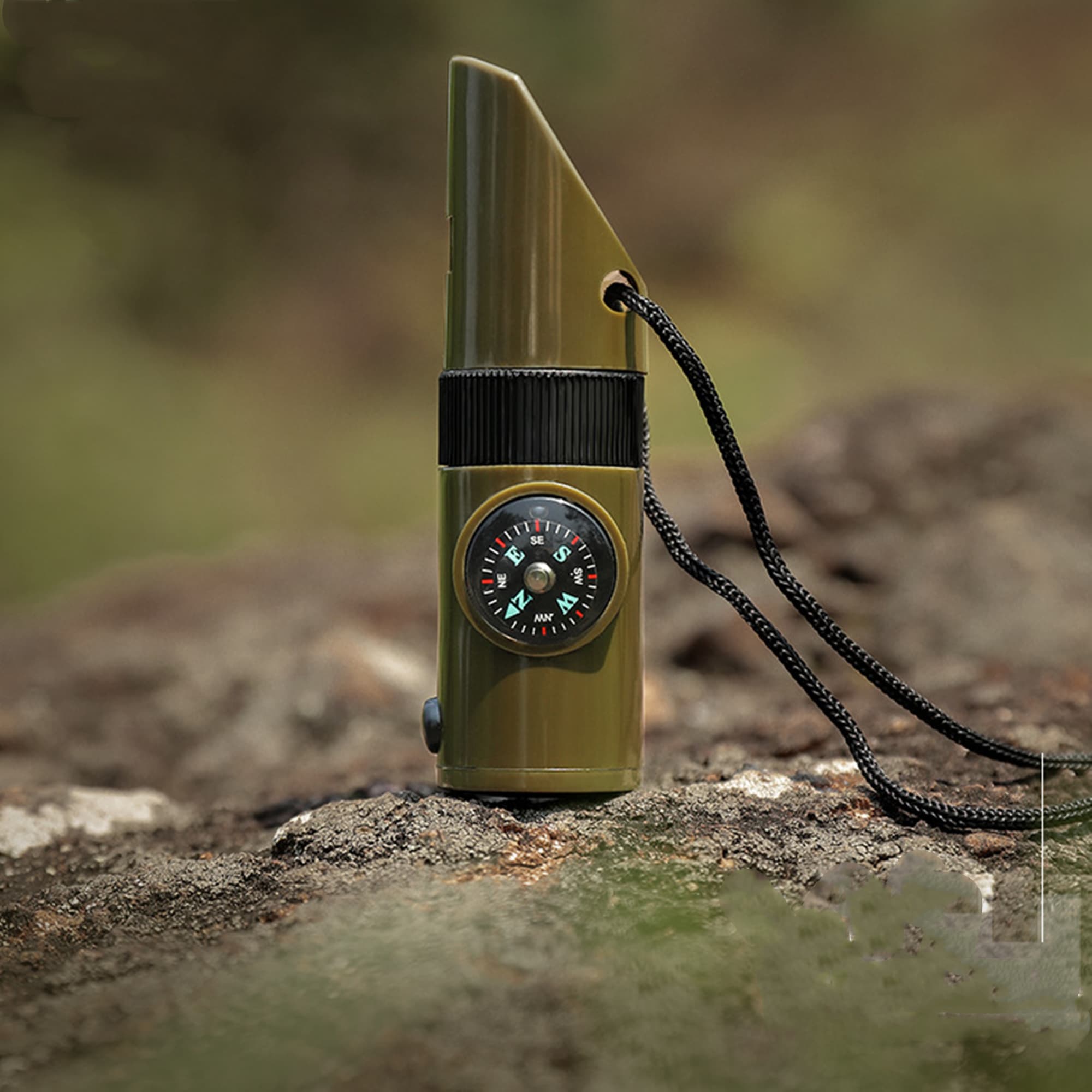 A green camping whistle with a built-in compass, attached to a black lanyard, resting on a rocky surface. A green camping whistle with a built-in compass, attached to a black lanyard, resting on a rocky surface.