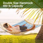 A double hammock in blue and white quilted fabric, suspended between trees near the ocean, with two people relaxing.