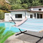 A striped hammock in blue, white, and green fabric displayed by a poolside, featuring a wooden support and a cushion.
