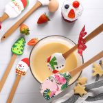A set of festive baking spatulas in red, green, and white, resting in a bowl of batter with assorted ingredients nearby.