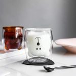 A cute bear cup in transparent glass with a white interior, featuring a playful bear design, on a black coaster.