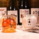 Two cute bear-shaped cups, one amber and the other white, sit on a countertop with wine bottles in the background.