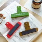 Three magnetic plastic clips in green, red, and brown lying on an open book, with a candle in the background.