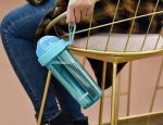A drink bottle in turquoise with a compartmentalised double drinking straw, held by a person sitting on a gold chair.