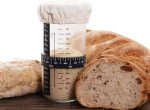 A transparent 32 oz. Mason fermenter jar with a cloth cover, surrounded by various types of freshly baked bread.