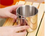 A stainless steel camping mug with a folding handle, displayed on a wooden surface. The mug is shiny and metallic.