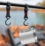 A stainless steel camping mug with a folding handle, suspended from hooks by straps against a blurred outdoor background.