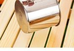 A stainless steel camping mug with a folding handle, resting on wooden planks. The mug has a shiny finish and a logo.