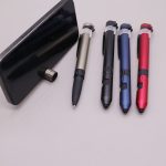 A smartphone with a phone stand beside four multi-tool pens in silver, black, blue, and red. Each has a logo.