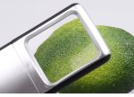 A silver slide-out magnifier with light focusing on a green fruit, likely an avocado. It features a logo.