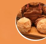 A plush brown teddy bear wearing a hoodie sits on a wooden surface. It has round, fluffy paws.