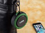 A green waterproof Bluetooth speaker hangs from a black bag, with a smartphone resting nearby. The speaker has a logo.